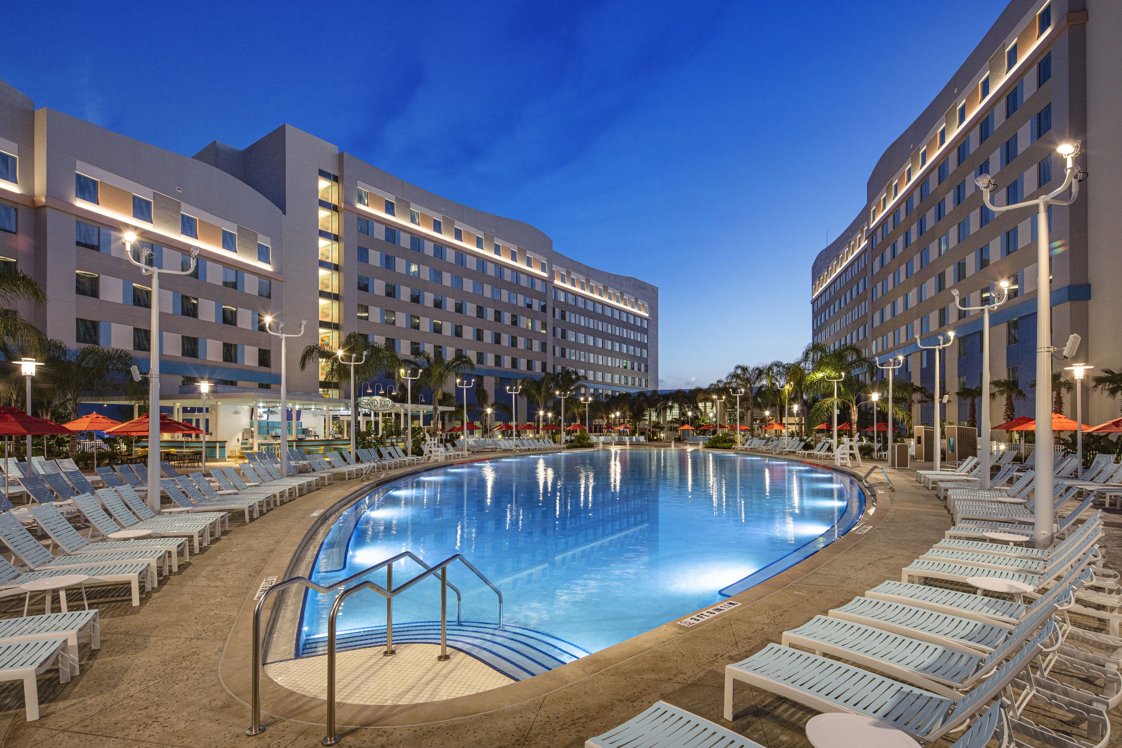 The image shows a modern hotel complex with a large swimming pool surrounded by lounge chairs. The scene is illuminated by evening lights, creating a tranquil atmosphere.