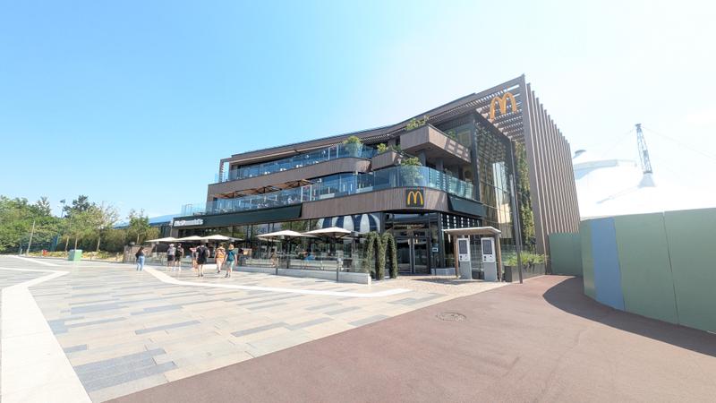 The image depicts a modern McDonald's restaurant with a sleek architectural design, featuring outdoor seating areas and greenery. The restaurant is located in a sunny environment, indicating a welcoming atmosphere for customers.