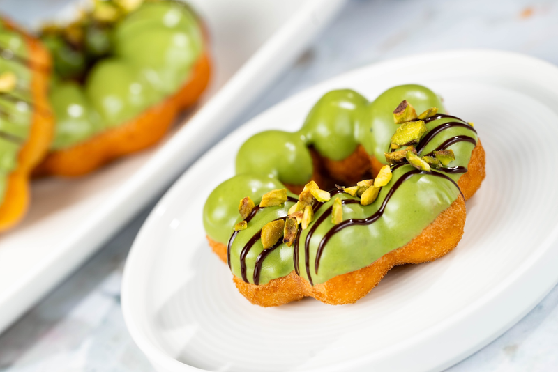 The image showcases a vibrant green doughnut topped with pistachios and a drizzle of chocolate, presented on a white plate. In the background, another similar doughnut can be seen, highlighting the dessert's appealing colors and textures.
