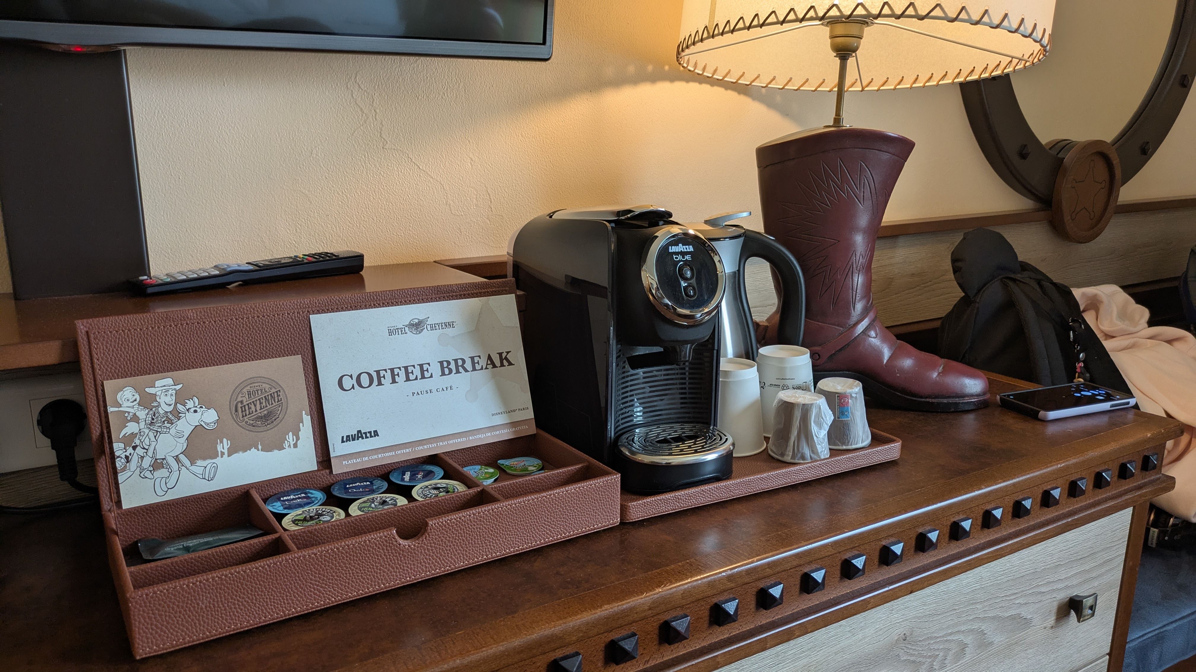 The image depicts a neatly arranged coffee station on a wooden table, featuring a coffee machine and various coffee pods in a decorative box. A leather boot and a lamp are also visible in the scene, suggesting a cozy and casual setting.