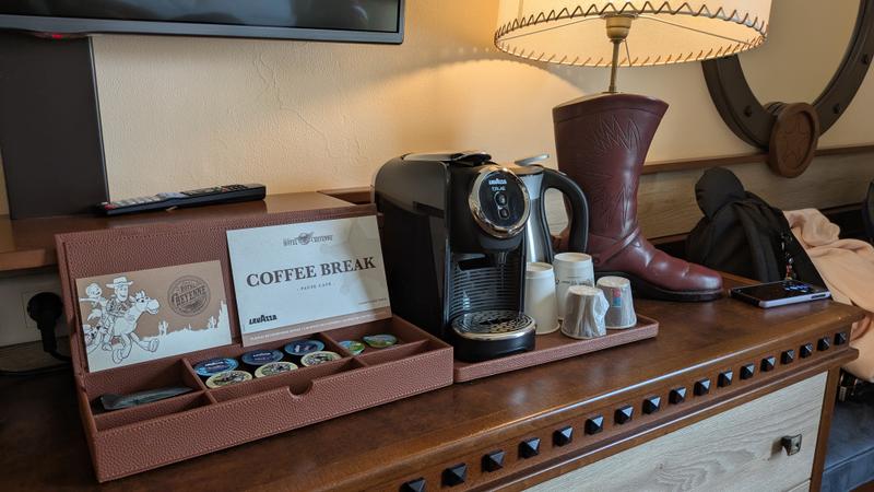 The image depicts a neatly arranged coffee station on a wooden table, featuring a coffee machine and various coffee pods in a decorative box. A leather boot and a lamp are also visible in the scene, suggesting a cozy and casual setting.