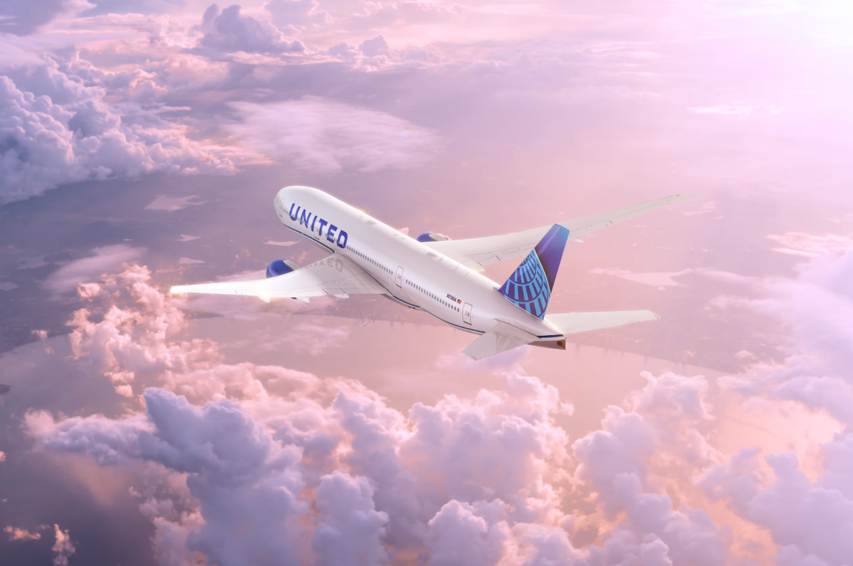 An airplane belonging to United Airlines is shown flying above a sea of clouds during a vibrant sunset. The aircraft is captured in mid-flight, showcasing its design against a colorful sky.