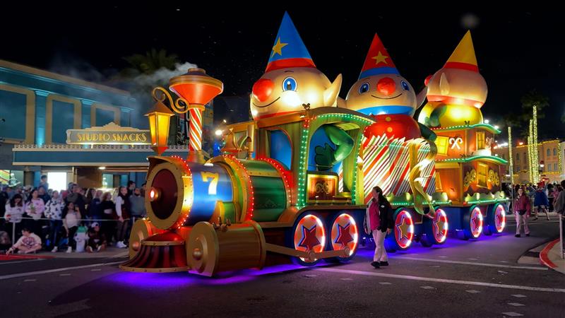 A brightly lit parade float featuring a colorful train with large clown figures at the front. The scene is set at night, surrounded by an audience gathered to watch the festivities.