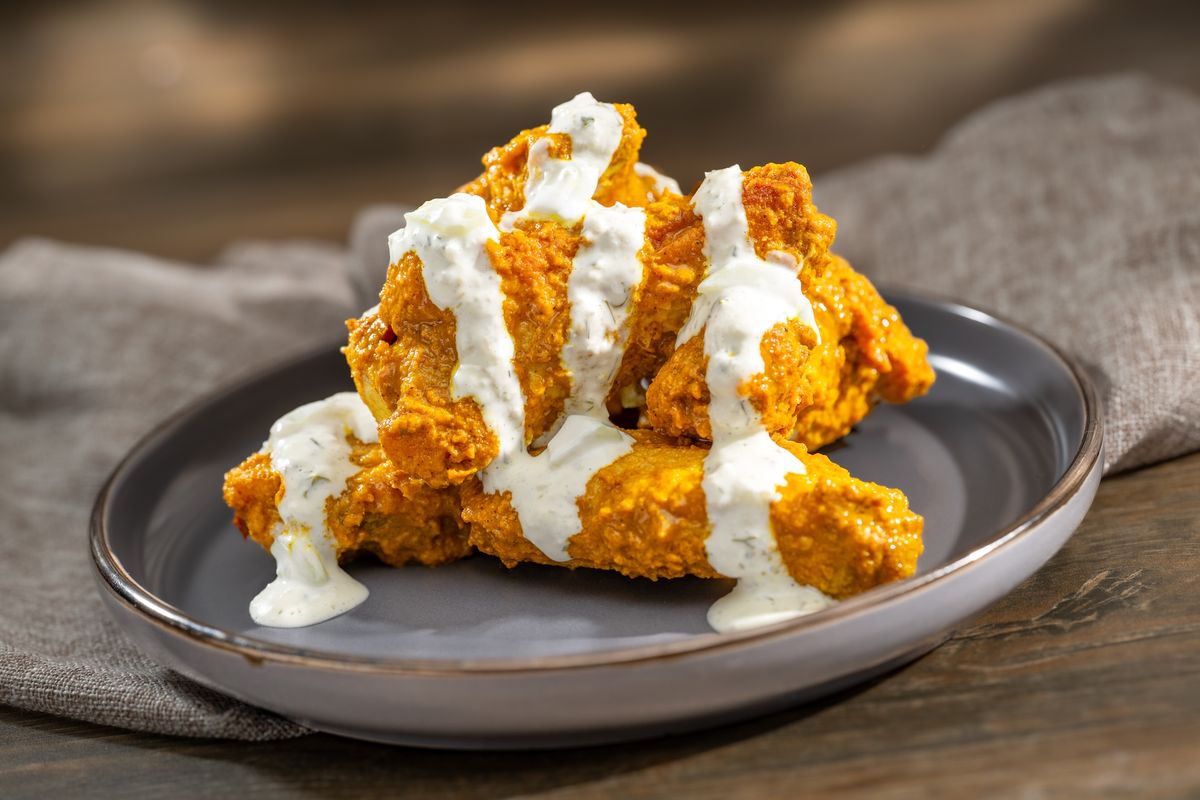 A close-up image of crispy chicken wings drizzled with a creamy sauce, presented on a gray plate. The wings are arranged on a textured surface, highlighting their golden-brown color and appetizing appearance.