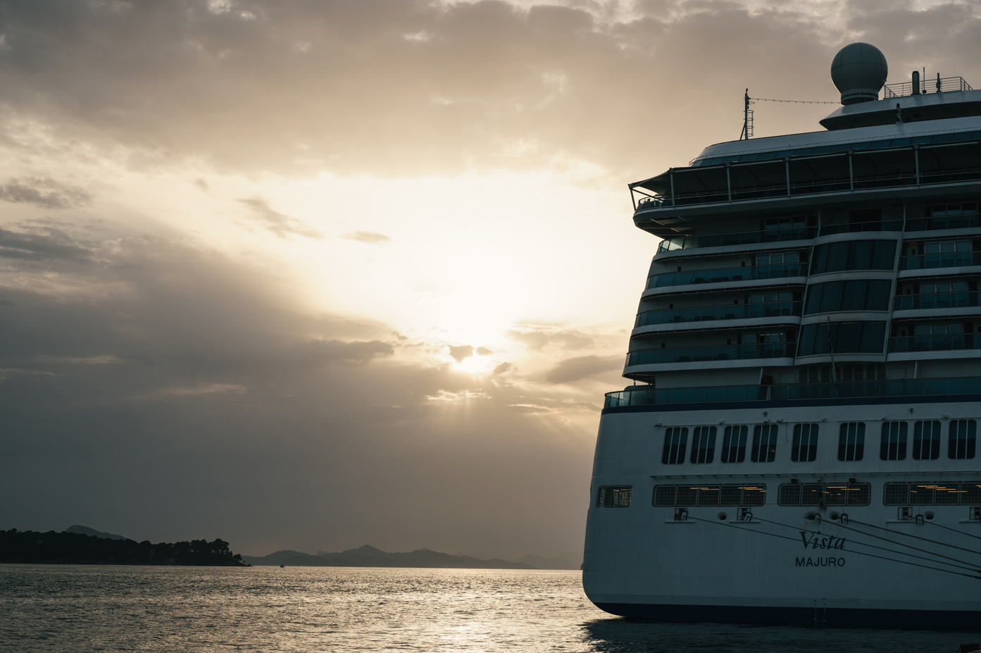 The image depicts a large cruise ship named 'Vista' alongside a calm body of water, silhouetted against a dramatic sunset. The sky features soft clouds illuminated by the fading sunlight, creating a serene and picturesque atmosphere.