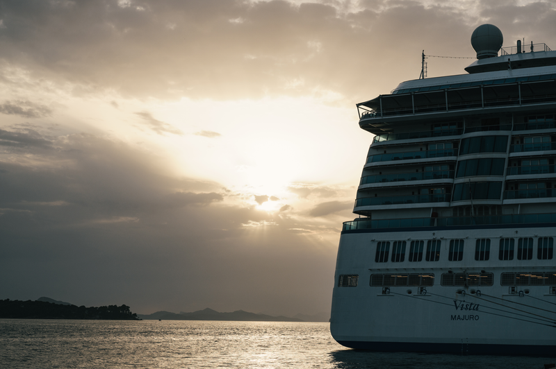 The image depicts a large cruise ship named 'Vista' alongside a calm body of water, silhouetted against a dramatic sunset. The sky features soft clouds illuminated by the fading sunlight, creating a serene and picturesque atmosphere.