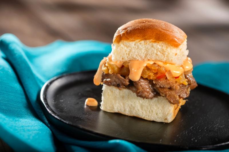 The image features a delicious pork slider on a black plate, topped with a creamy sauce and garnished with diced vegetables. The slider is positioned on a textured surface, complemented by a vibrant turquoise fabric in the background.