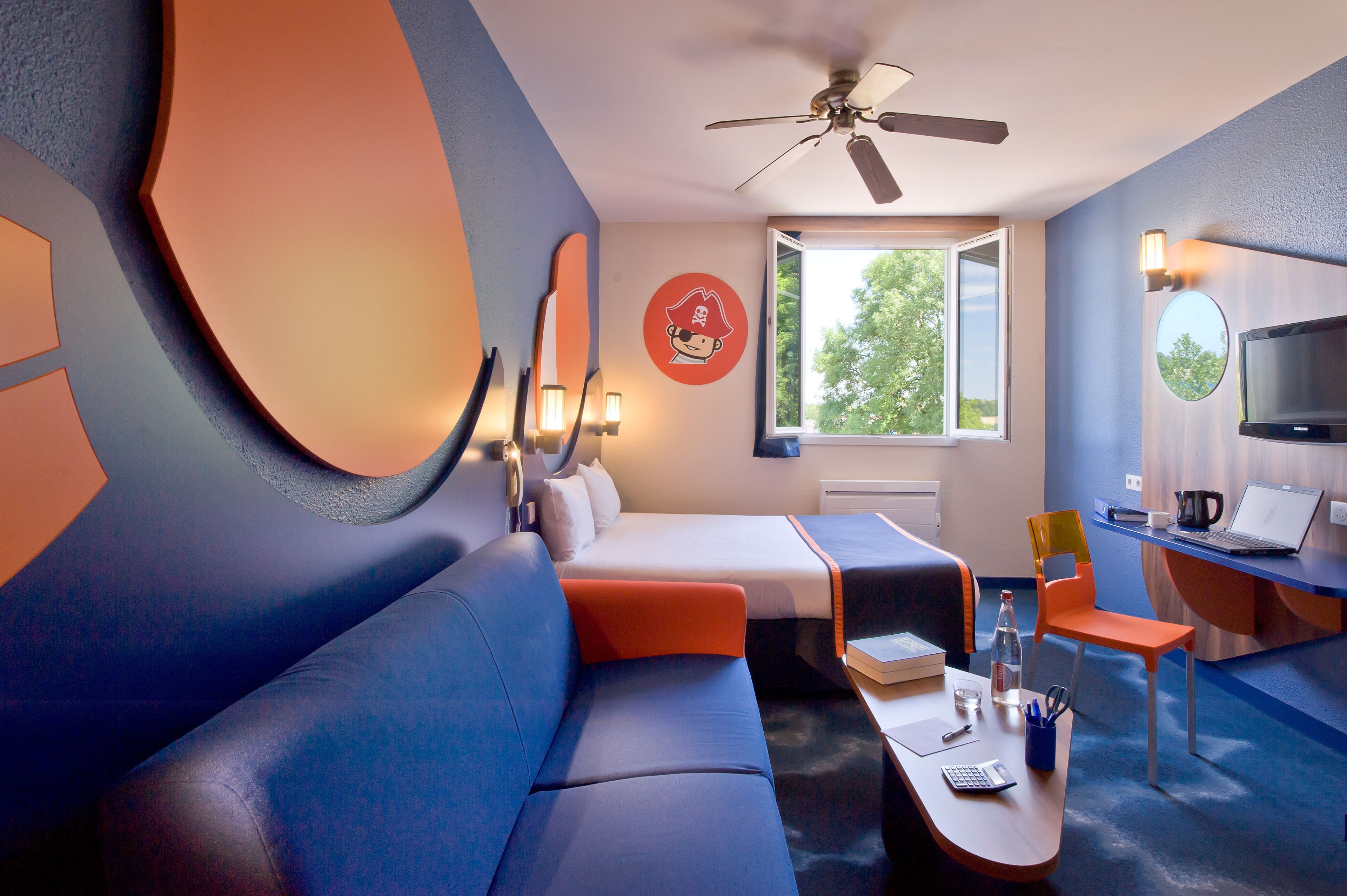 The image depicts a modern hotel room featuring a vibrant color scheme of blue and orange. It includes a bed, a sofa, a small desk, and a window with a view of greenery outside.