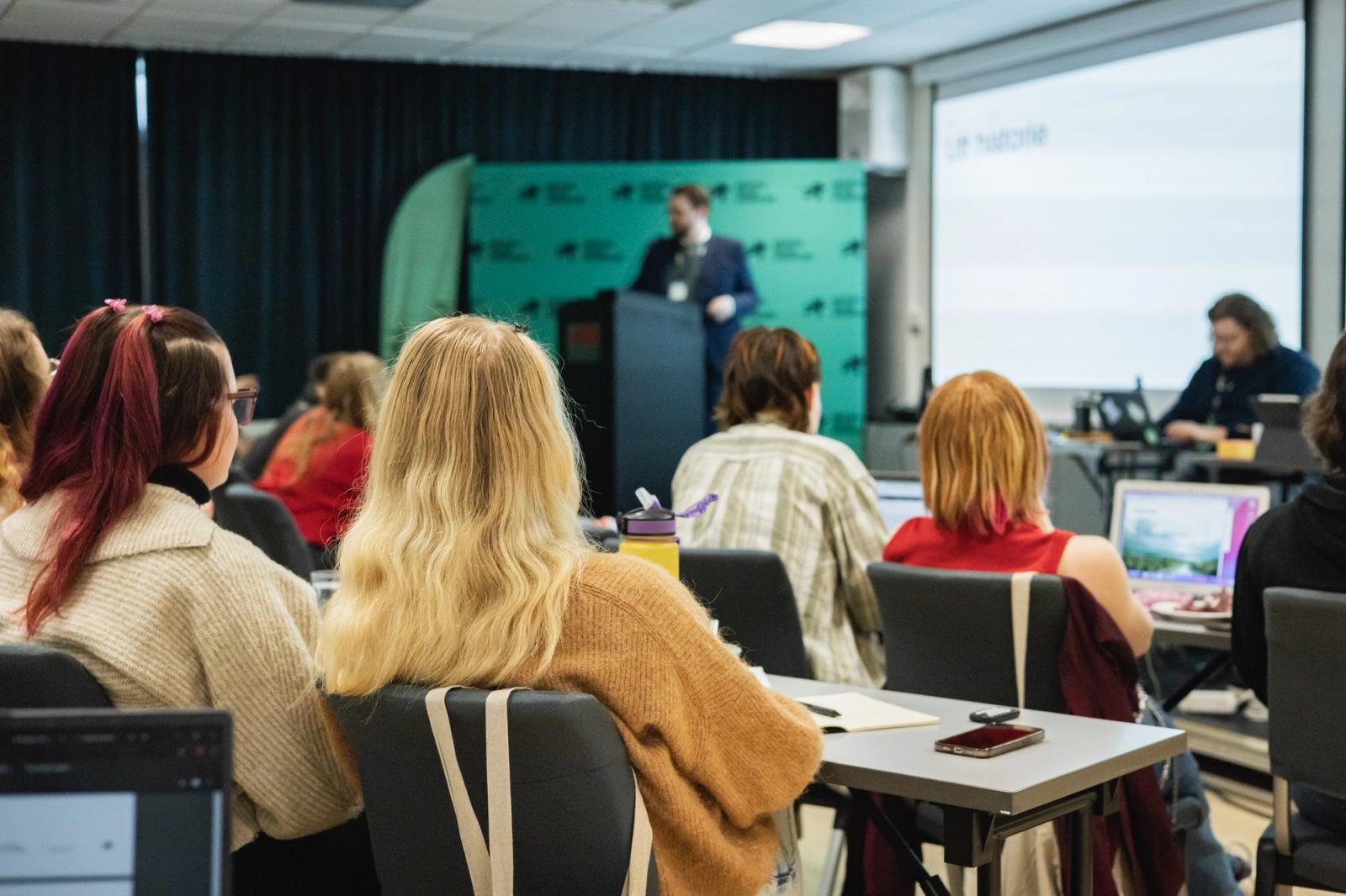 Bilde av person på talerstol foran en gruppe mennesker som hører på