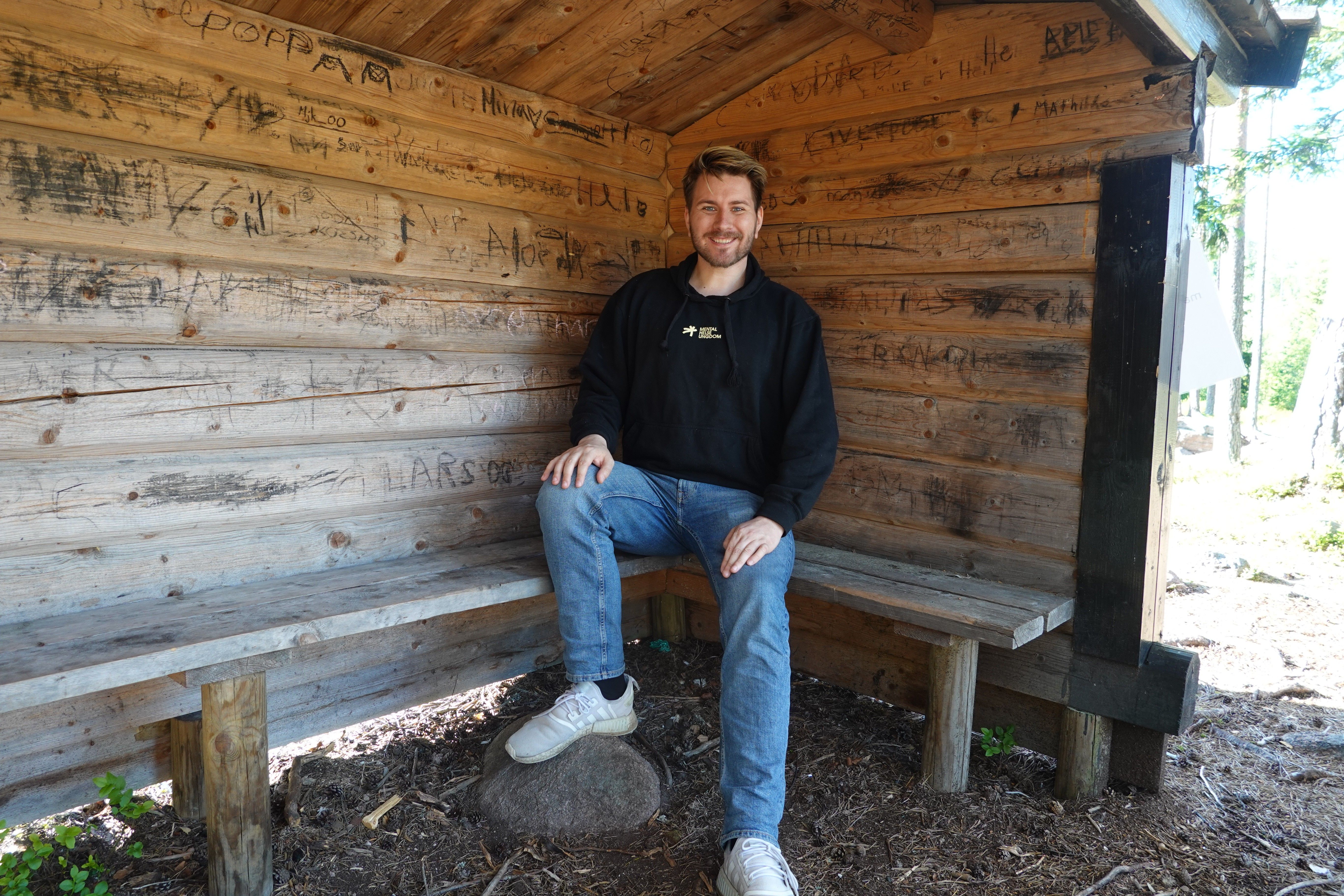 Christoffer sitter i et skur i skogen, med Mental Helse Ungdom-genser. Ser smilende i kamera