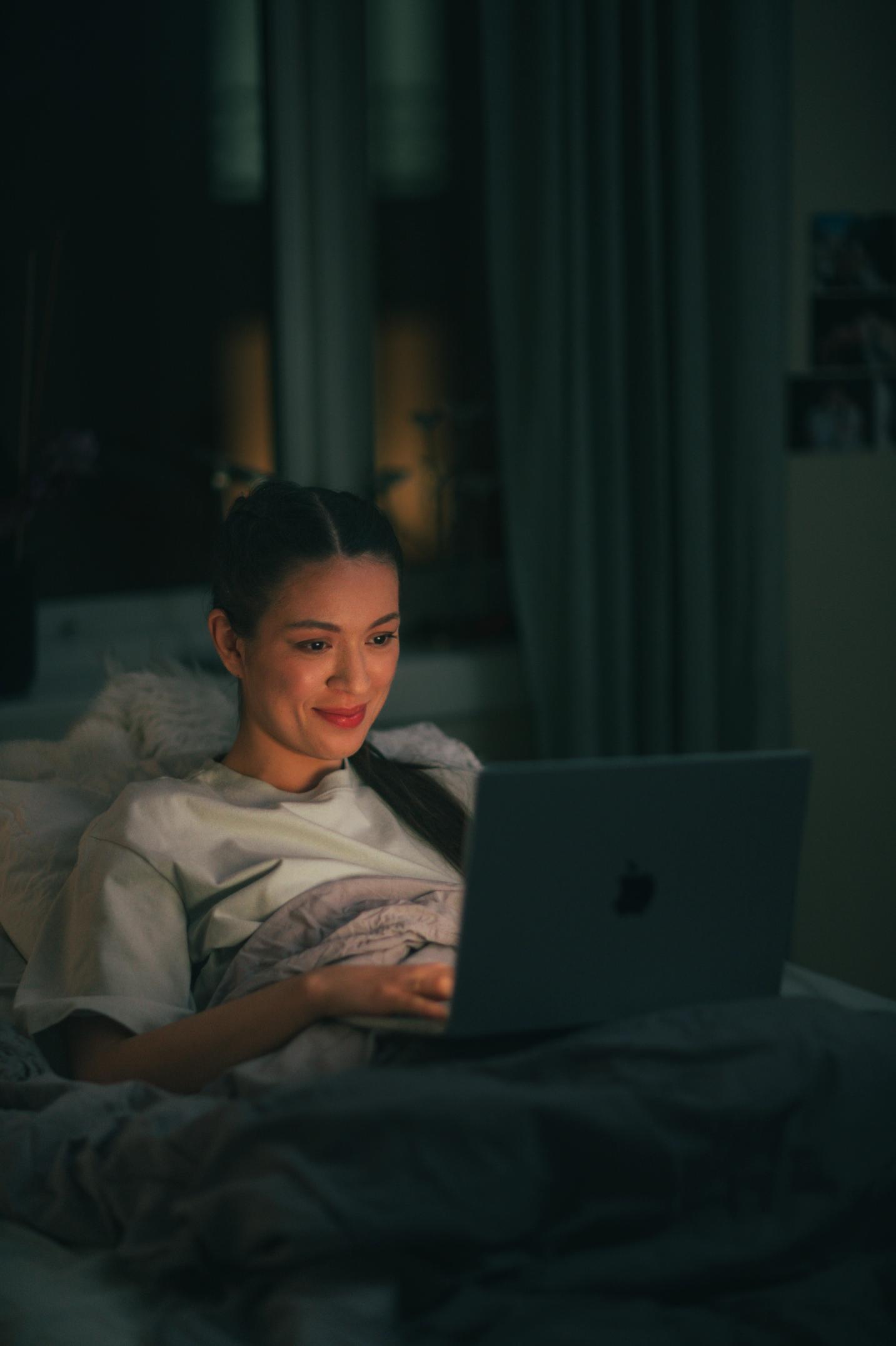 Kvinne som ligger i senga og ser på en åpen laptop.