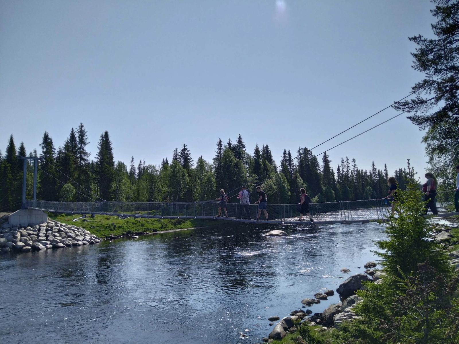 Deltakere på tur går på en bro over en elv i sommersol