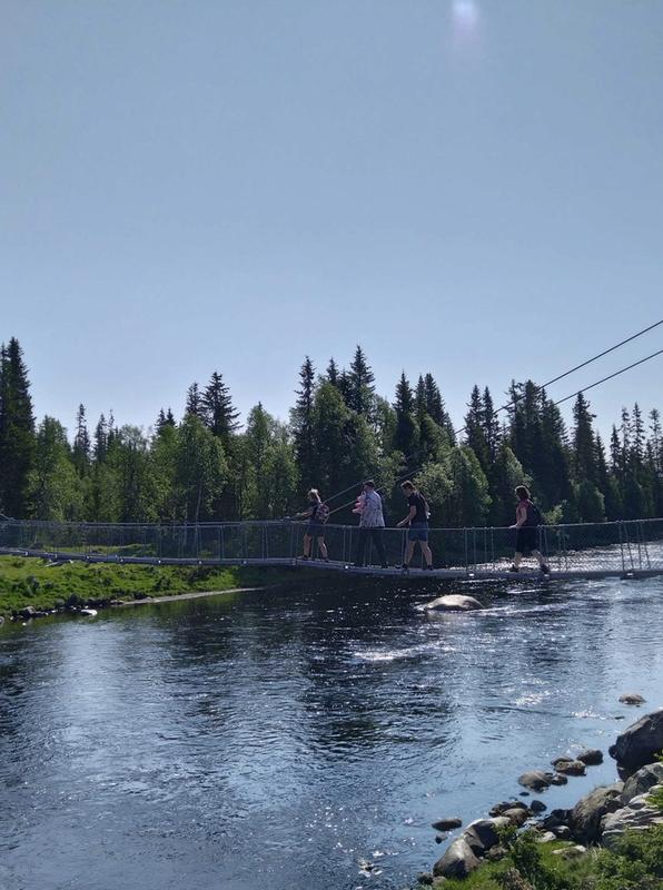 Deltakere på tur går på en bro over en elv i sommersol