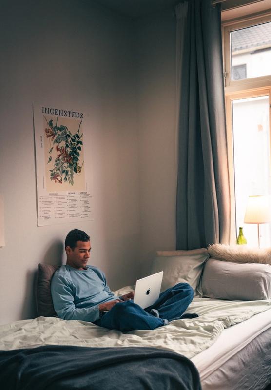 Ung mann sitter i sengen med laptop og smiler