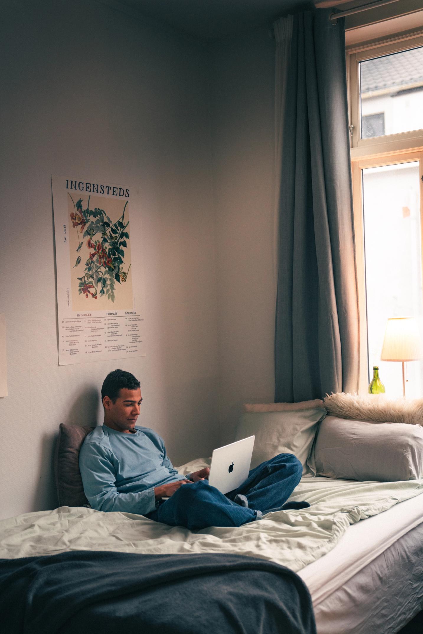 Ung mann sitter i sengen med laptop og smiler