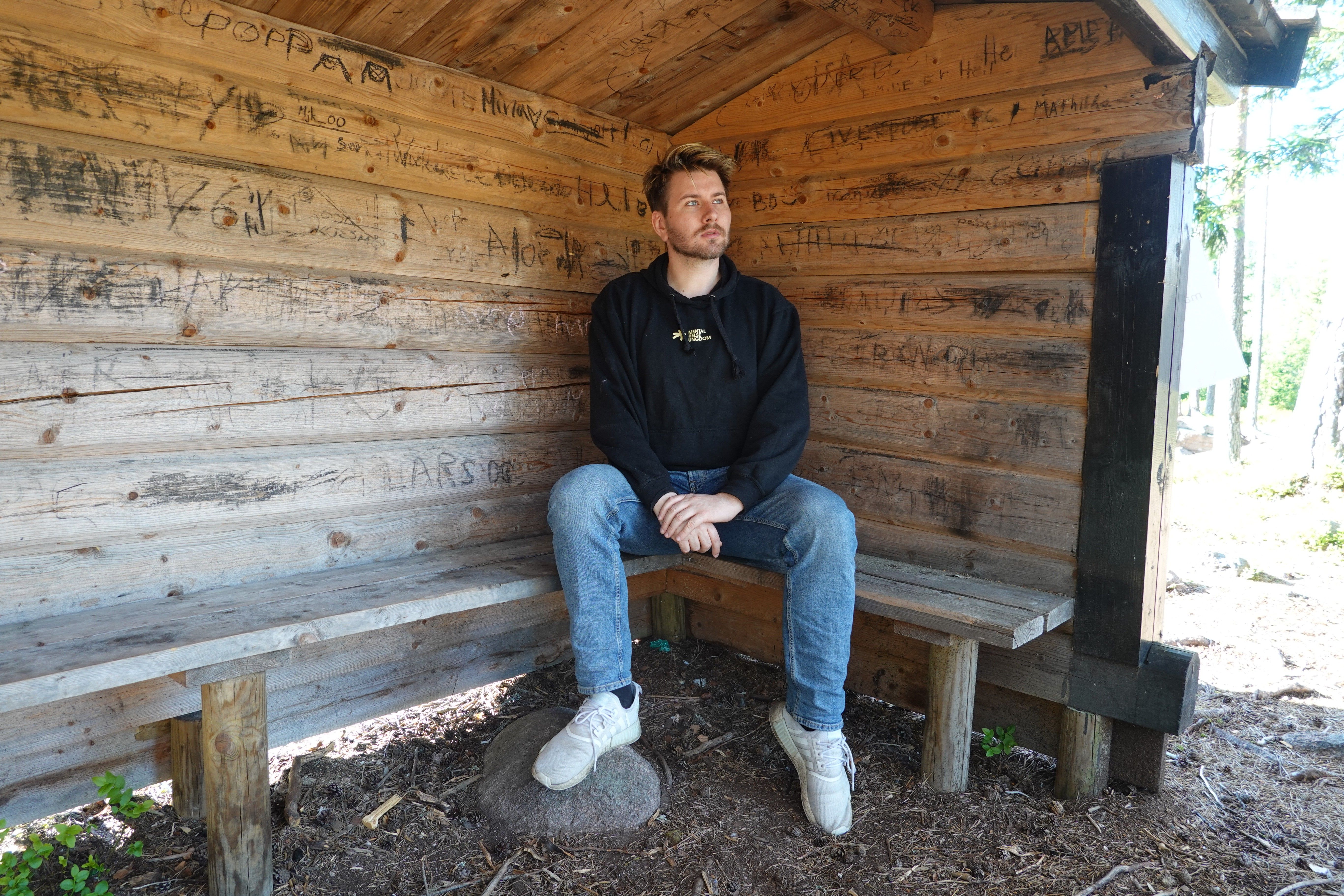 Christoffer sitter i et skur i skogen, med Mental Helse Ungdom-genser. Ser utover
