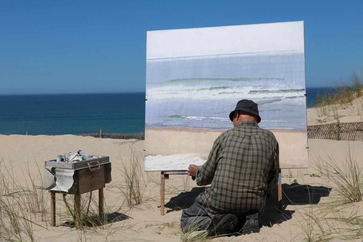 a man is kneeling down on the beach painting a picture of the ocean .