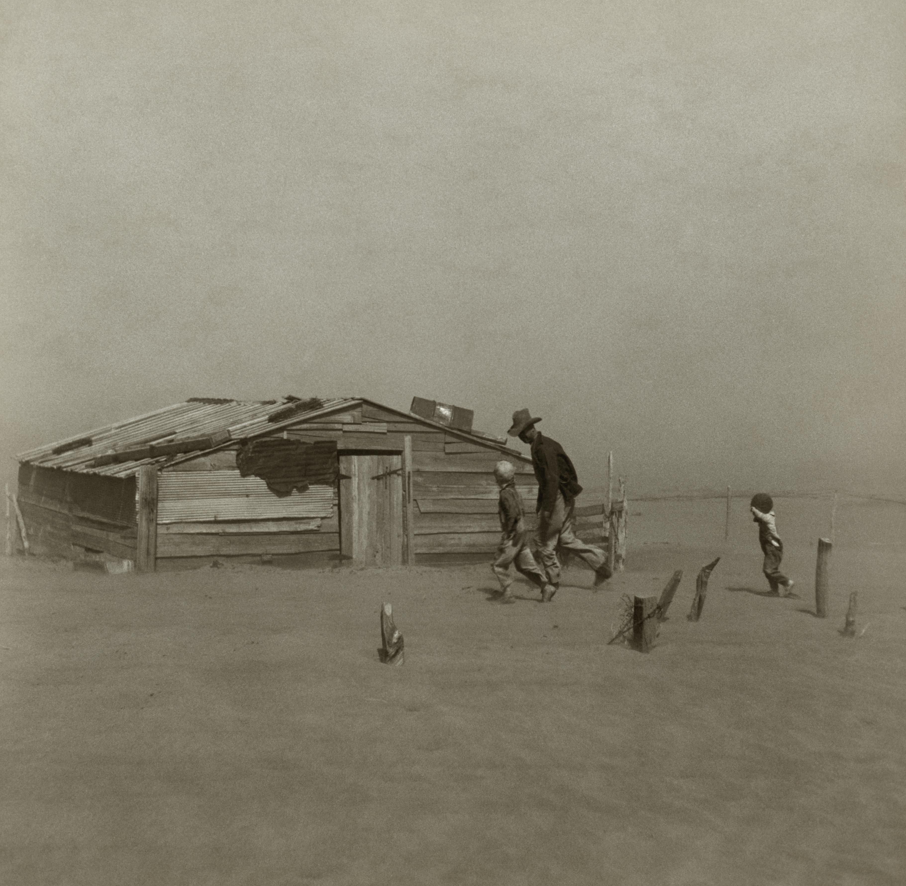 a black and white photo of a man and two children walking in front of a small wooden house .