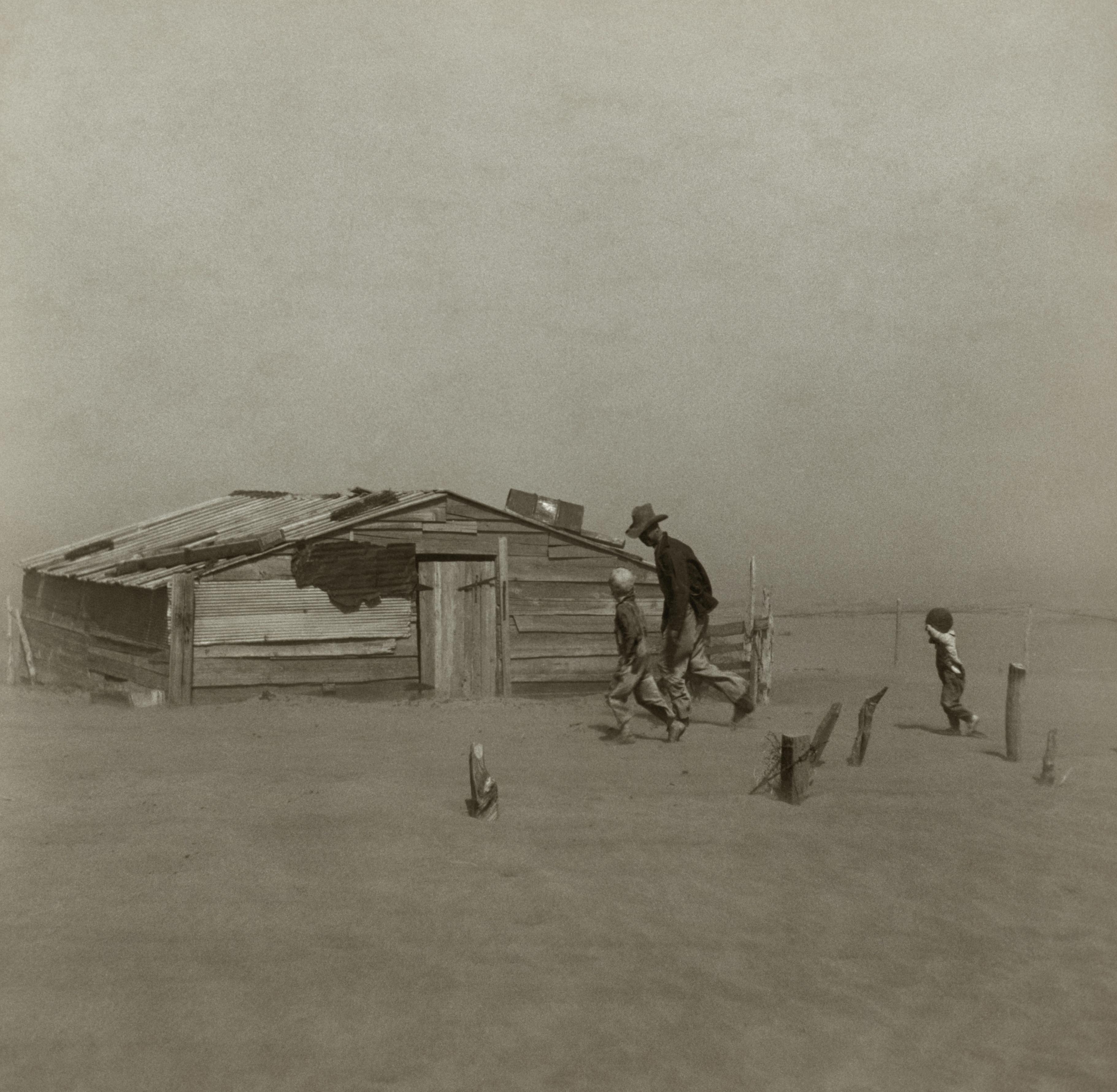 a black and white photo of a man and two children walking in front of a small wooden house .