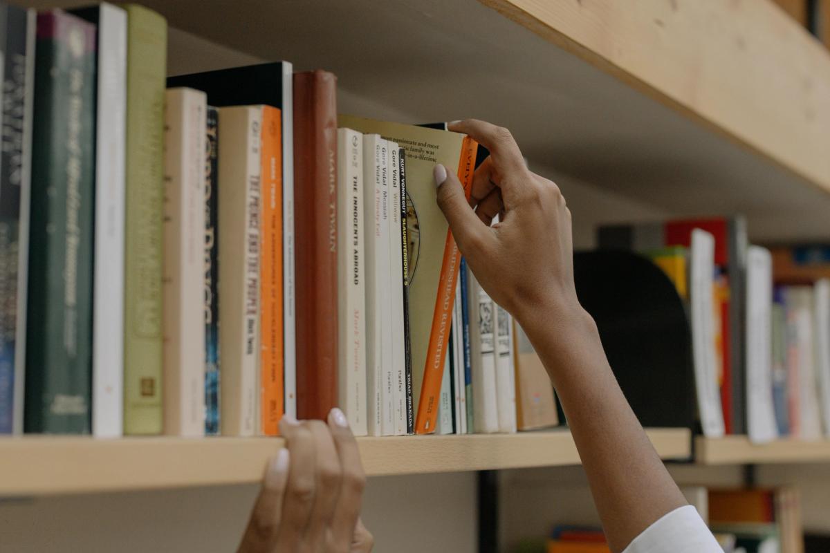 Person Holding A Book On Wooden Shelf Person Holding A Book On Wooden Shelf
