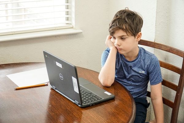 Boy sits in front of laptop computer appearing bored or disengaged. Text answer when a tutor is beneficial for a young student. 