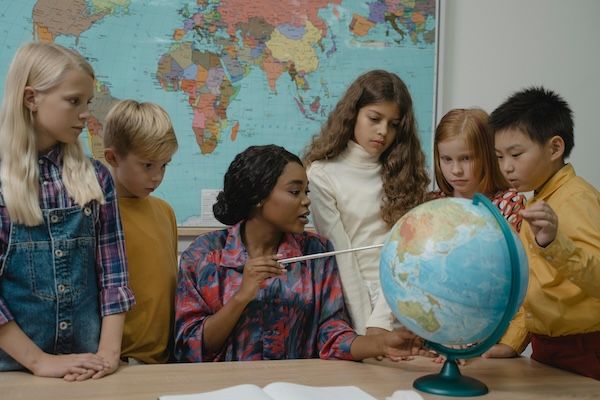 Photo depicts 5 students standing around a teacher's desk where she is pointing at a globe. Gifted kids are very curious about topics that interest them. 