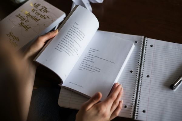 Hands holding an open book of what appears to be poetry. Book is resting on an open notebook. Hands holding an open book of what appears to be poetry. Book is resting on an open notebook.