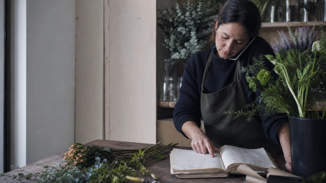 A florist cradles a phone to her ear while tracing a finger down a page of an order book at a workbench.