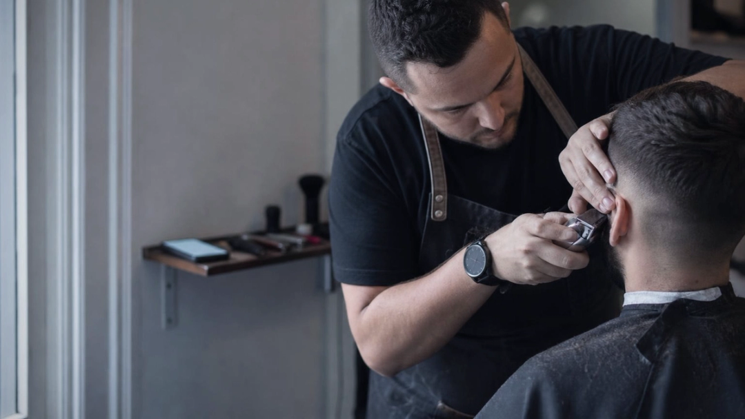 A barber in a dark apron trims the hairline behind a seated client's ear with clippers.