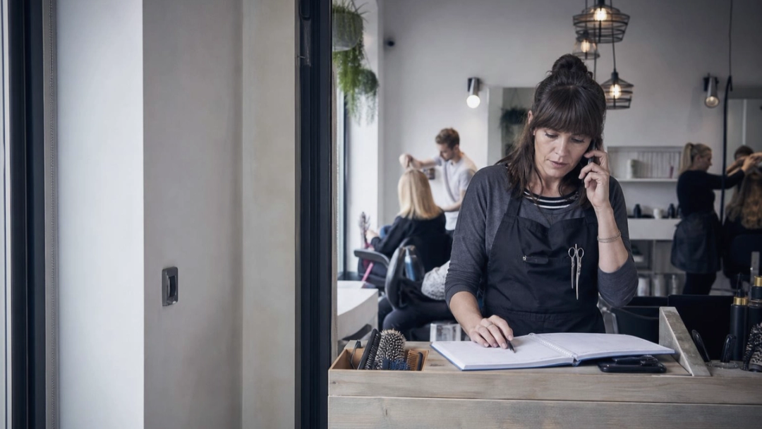Salon worker in an apron takes a phone call at the reception counter while checking an open appointment book.