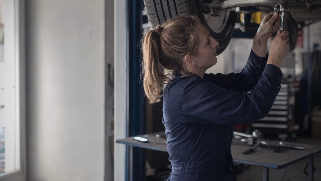 Mechanic in navy overalls reaches up with a socket wrench to a bolt under a car raised on a ramp.