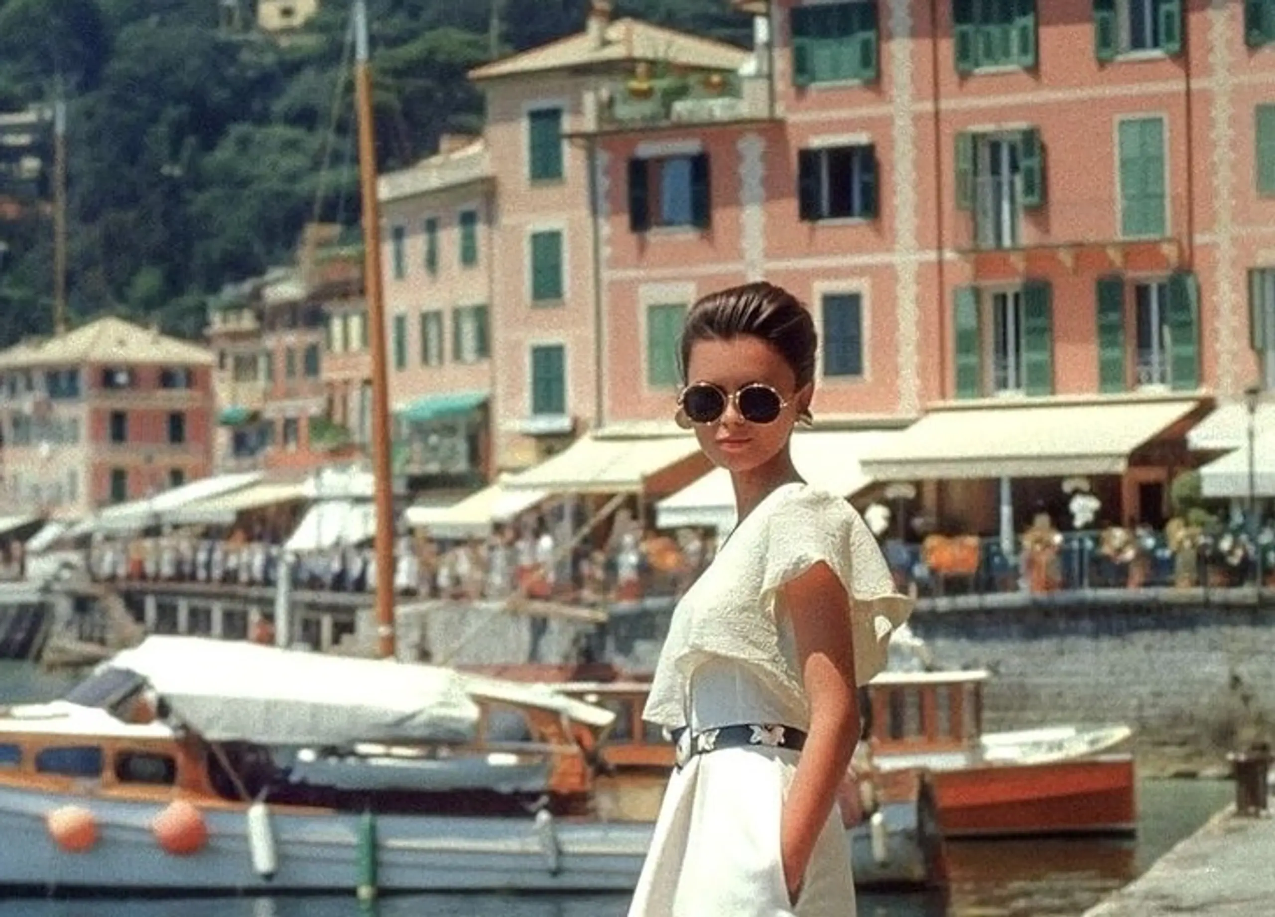 Woman in sunglasses standing by the Saint-Tropez harbor with colorful waterfront buildings and classic wooden boats