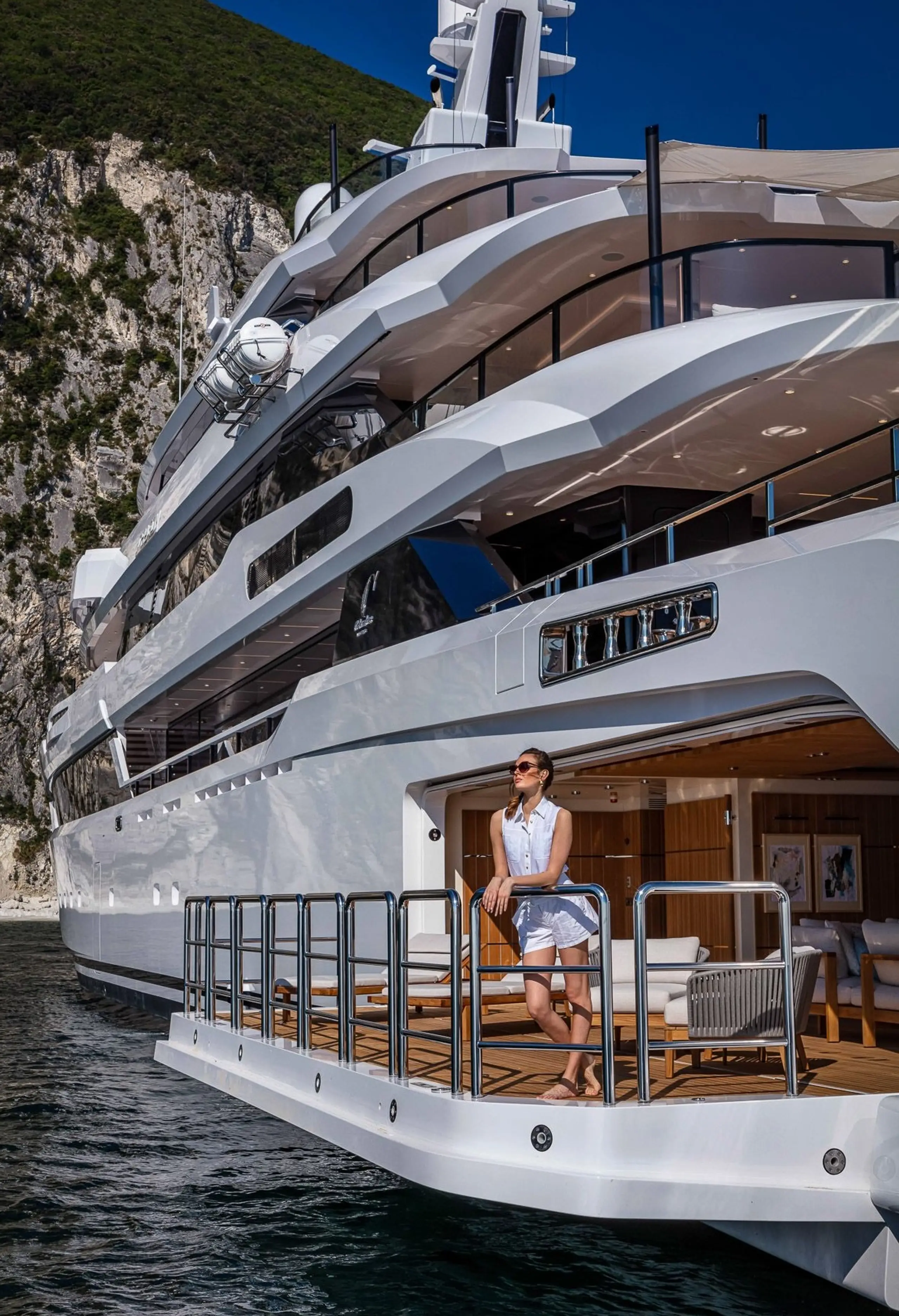 Guest standing on the aft deck of a superyacht near a rocky coastline.