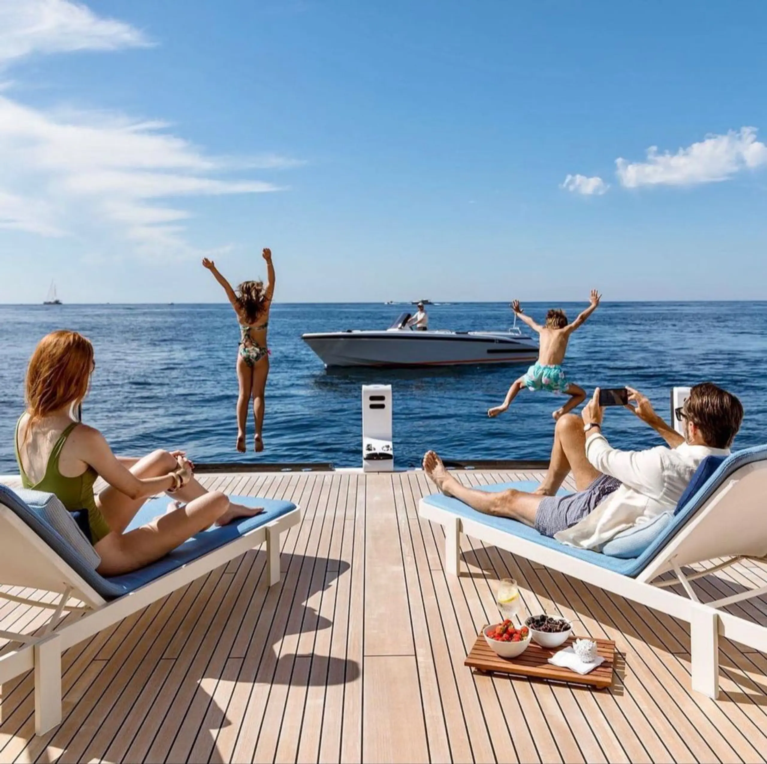 Family relaxing on a yacht deck while children jump into the sea.
