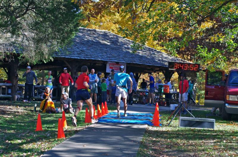 Runners finishing on the path at The Oval, past the race clock and the crowd under the shelter.