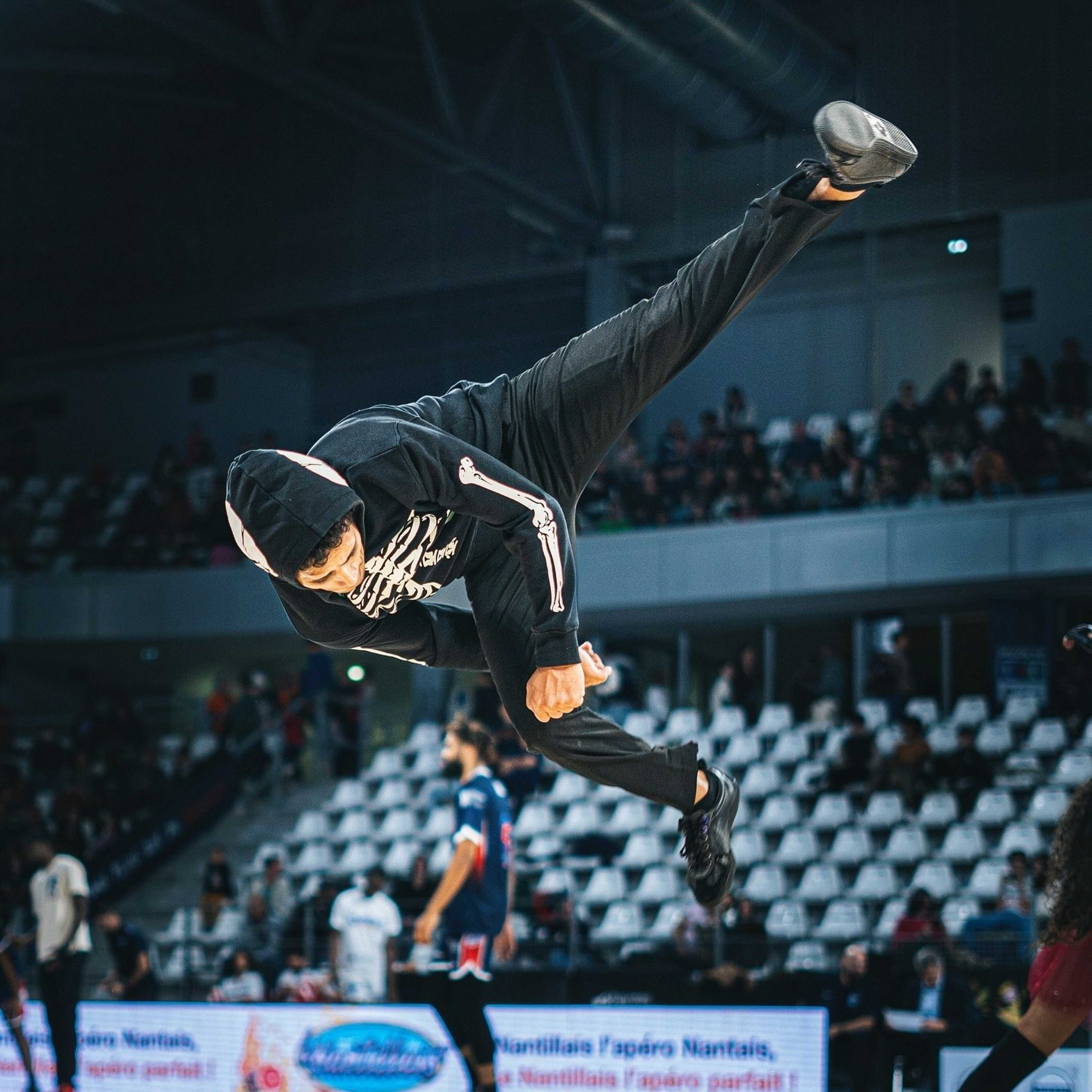 Acrobate en vrille horizontale en survêtement noir, tribunes en arrière-plan