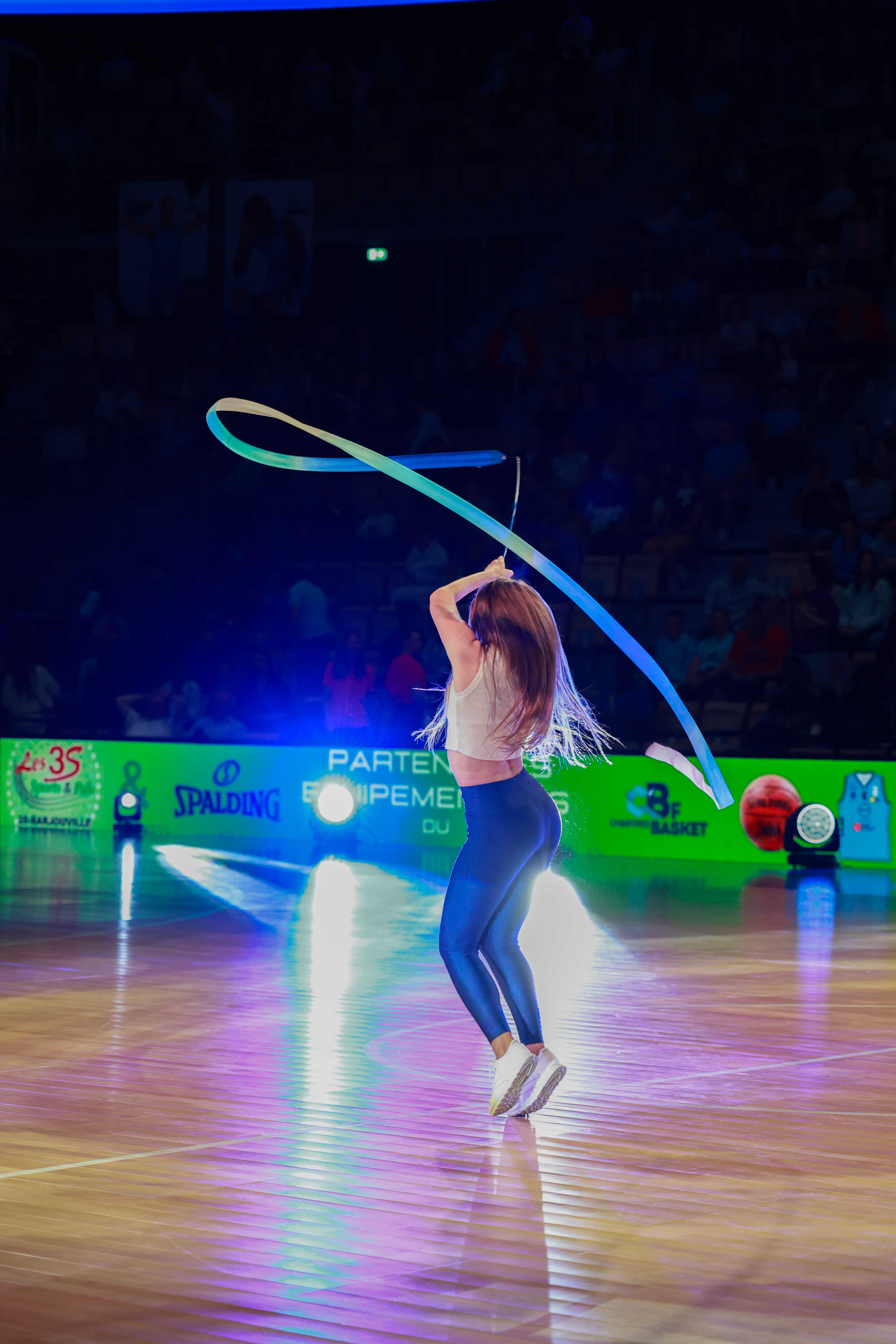 Danseuse de dos traçant une grande boucle de ruban turquoise sur un parquet de basket