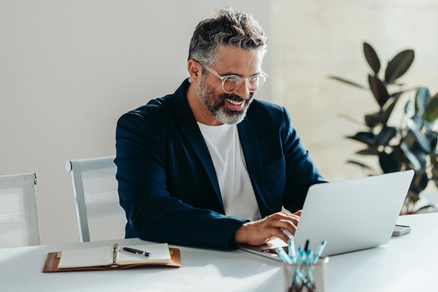 Cheerful, mature businessman with a beard performs enterprise seo optimisation on his laptop at a tidy desk in a modern office.