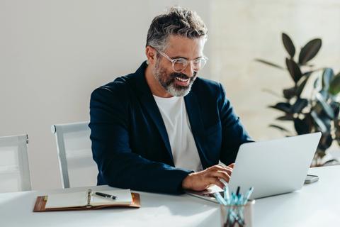 Cheerful, mature businessman with a beard performs enterprise seo optimisation on his laptop at a tidy desk in a modern office.