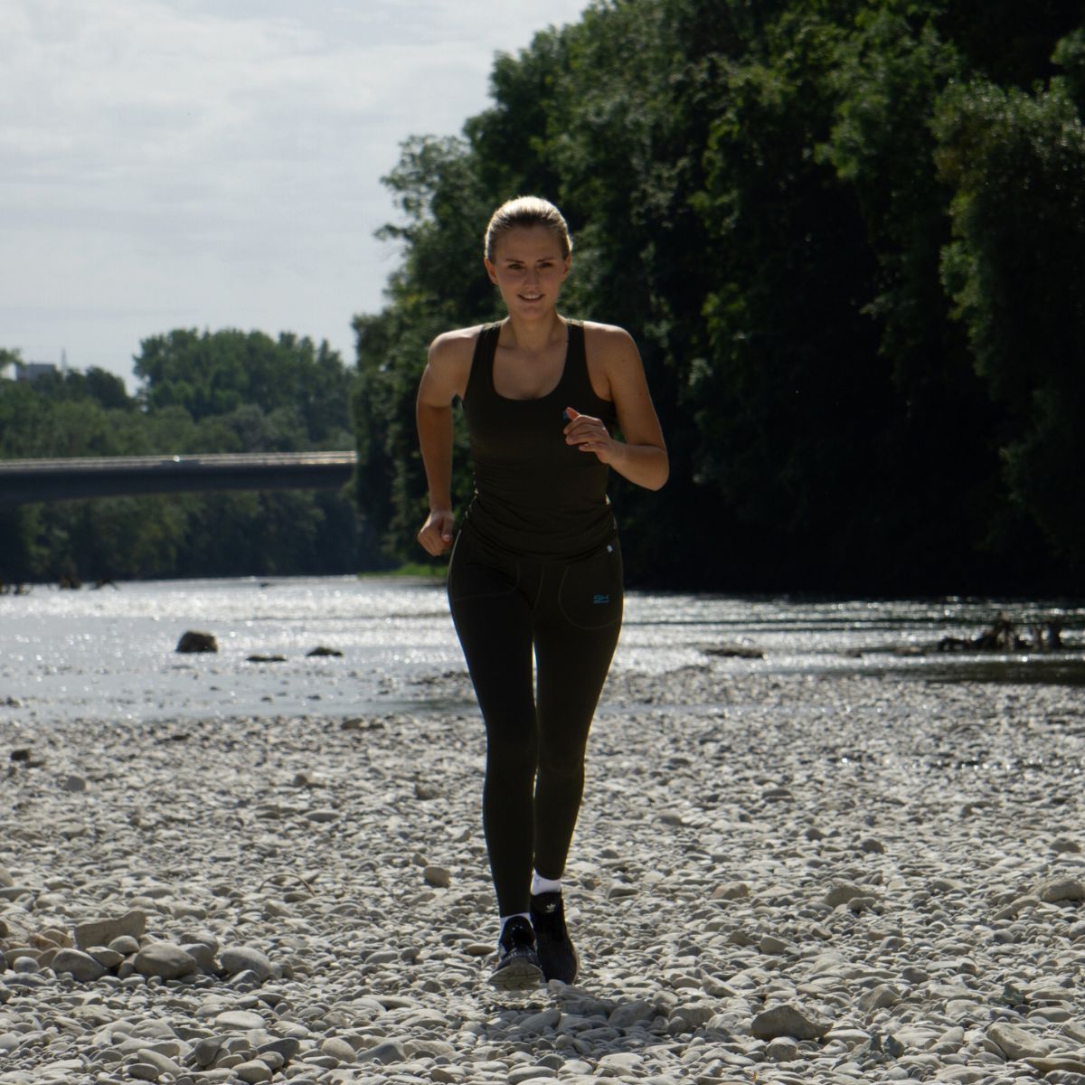 Frau im khaki farbnen Tennis Outfit von SPORTKIND, joggt energisch am Fluss 