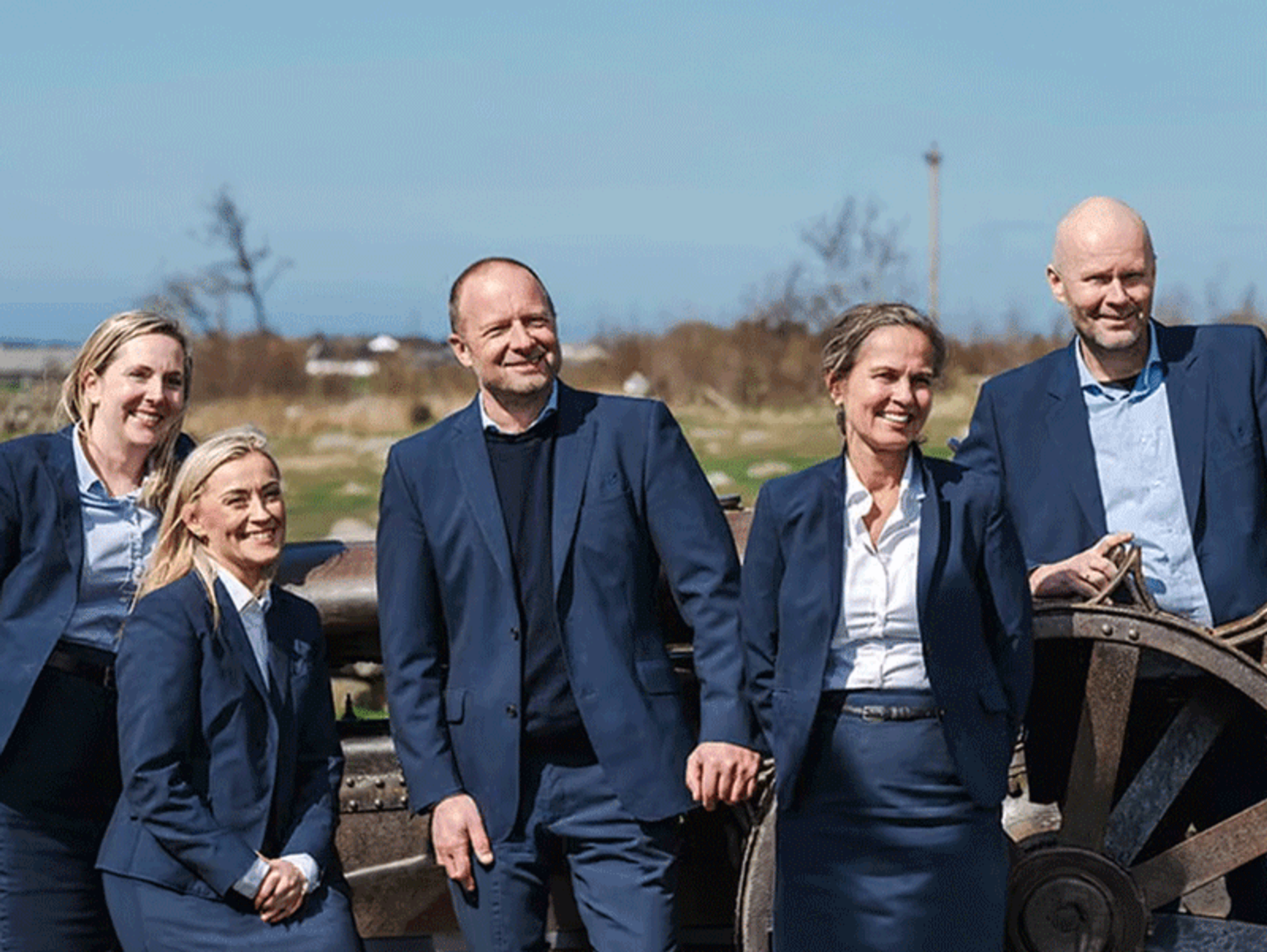 Fem smilende personer i mørke blå dresser står utendørs foran et gammelt hjul, med åpent landskap og gårdsbygninger i bakgrunnen.