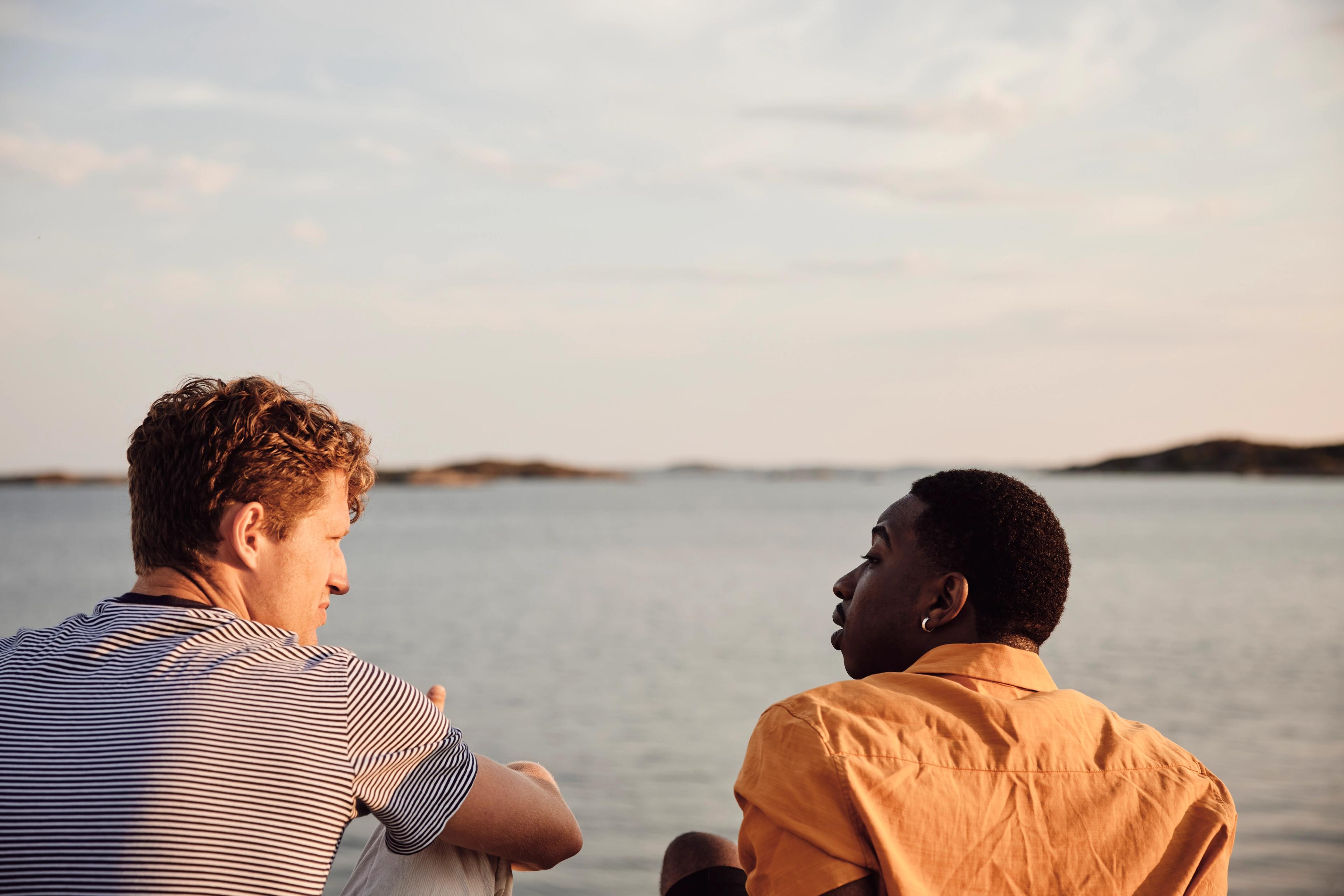 To personer sitter ved sjøen i varmt kveldslys og snakker sammen, med lave øyer i bakgrunnen.