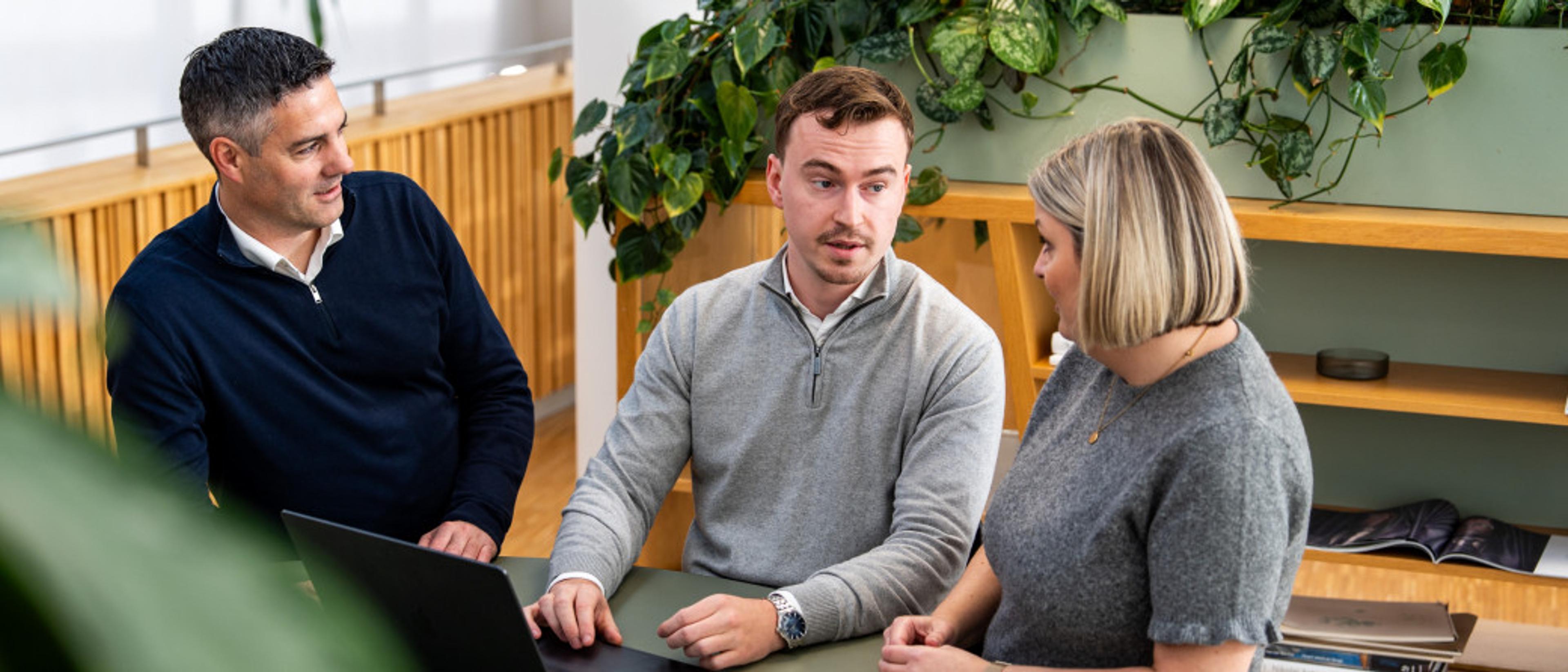 Tre kolleger sitter rundt et bord og diskuterer mens de arbeider ved en bærbar datamaskin i et lyst kontormiljø med grønne planter.