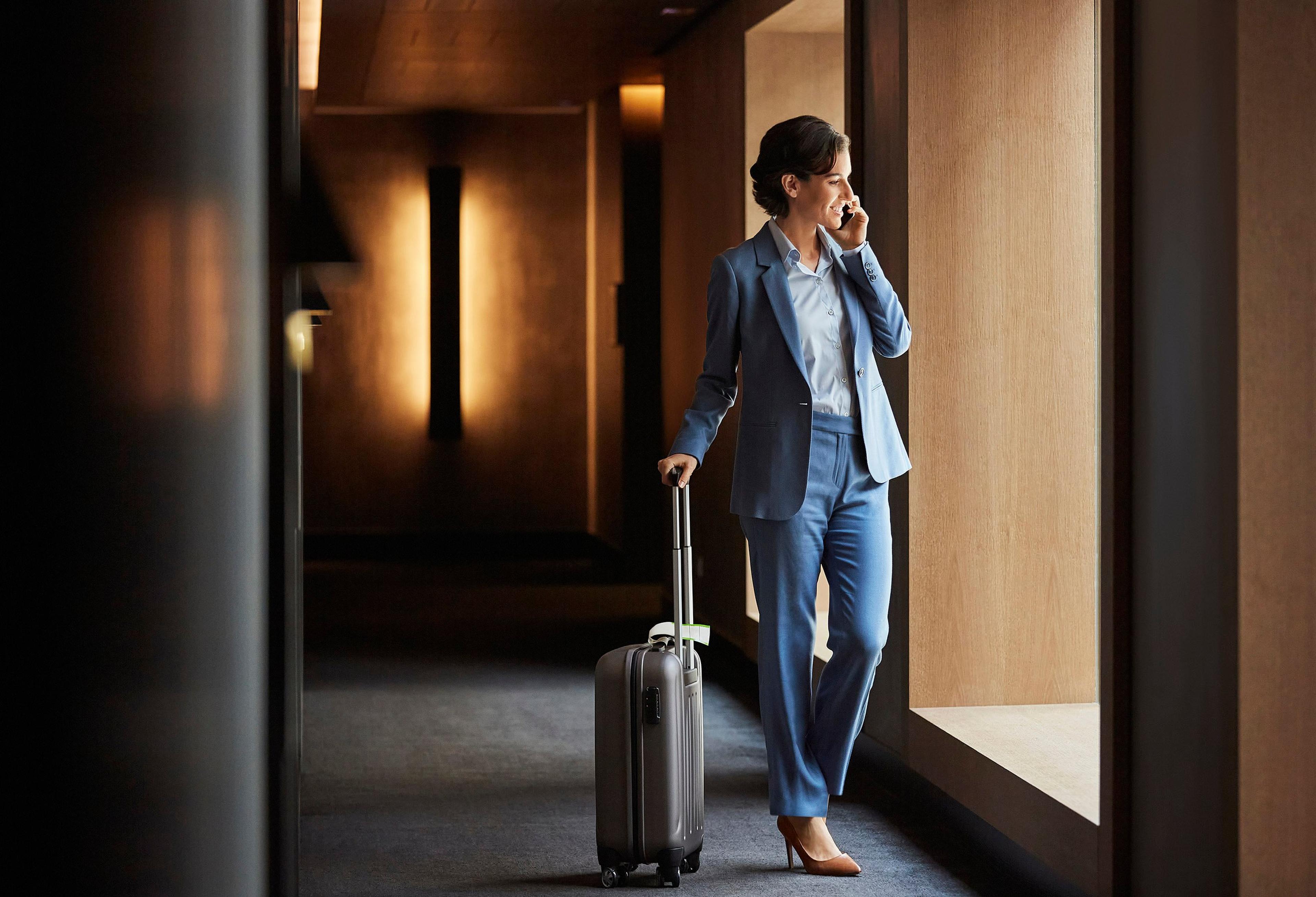 En forretningskvinne i blå dress snakker i mobiltelefon mens hun går gjennom en hotellkorridor med en trillekoffert.