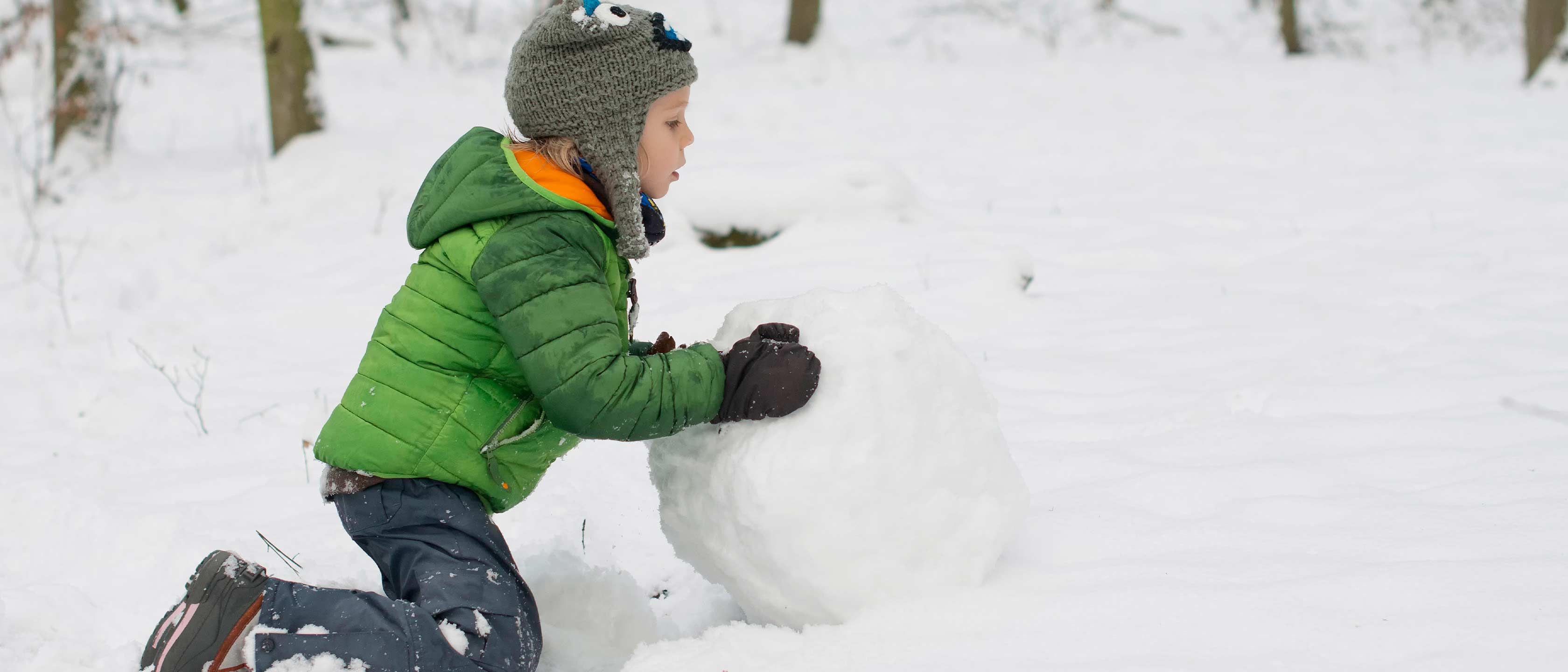 Et barn i grønn vinterjakke og grå lue kneler i snøen og ruller en stor snøball i en snødekt skog.