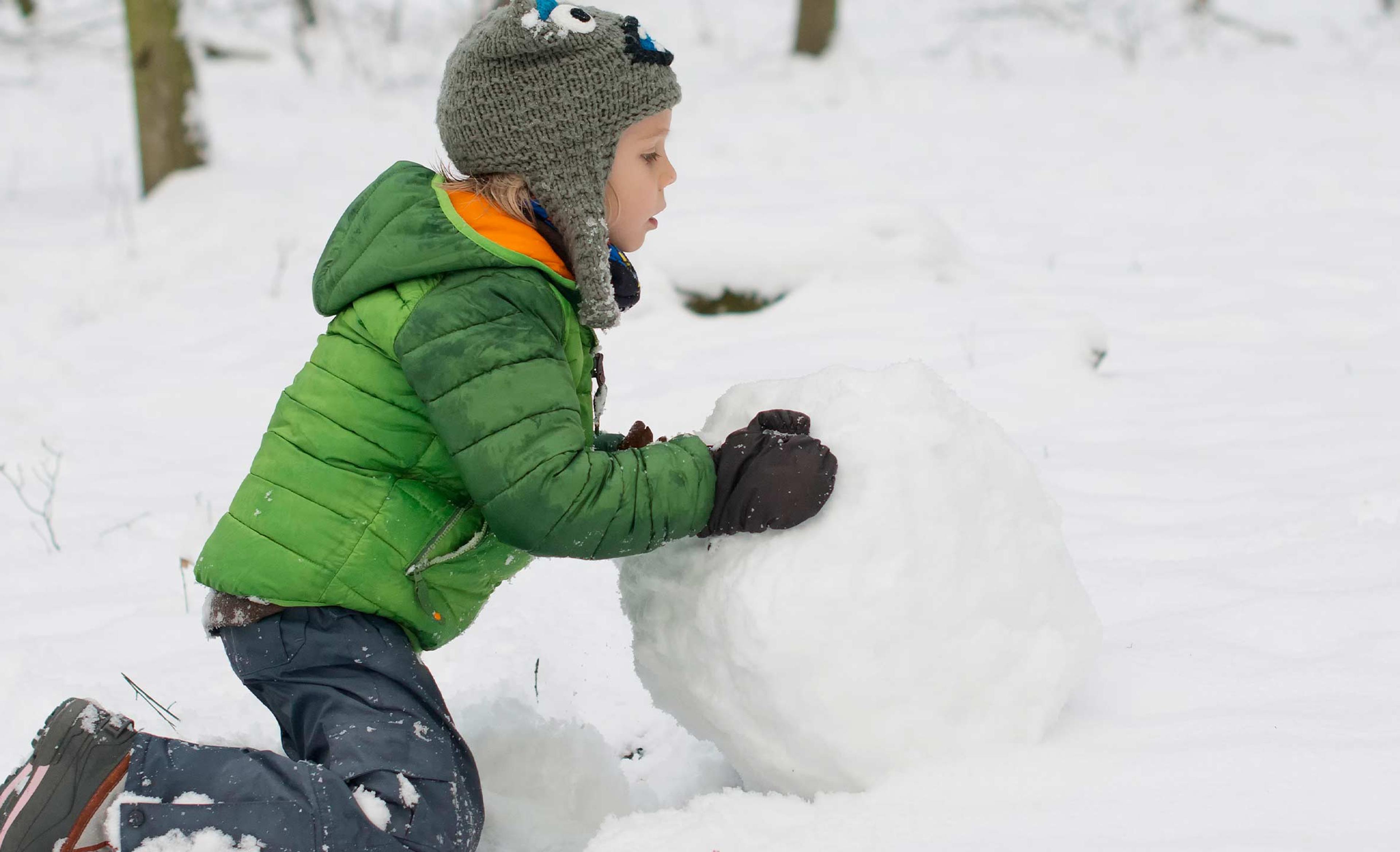 Et barn i grønn vinterjakke og grå lue kneler i snøen og ruller en stor snøball i en snødekt skog.