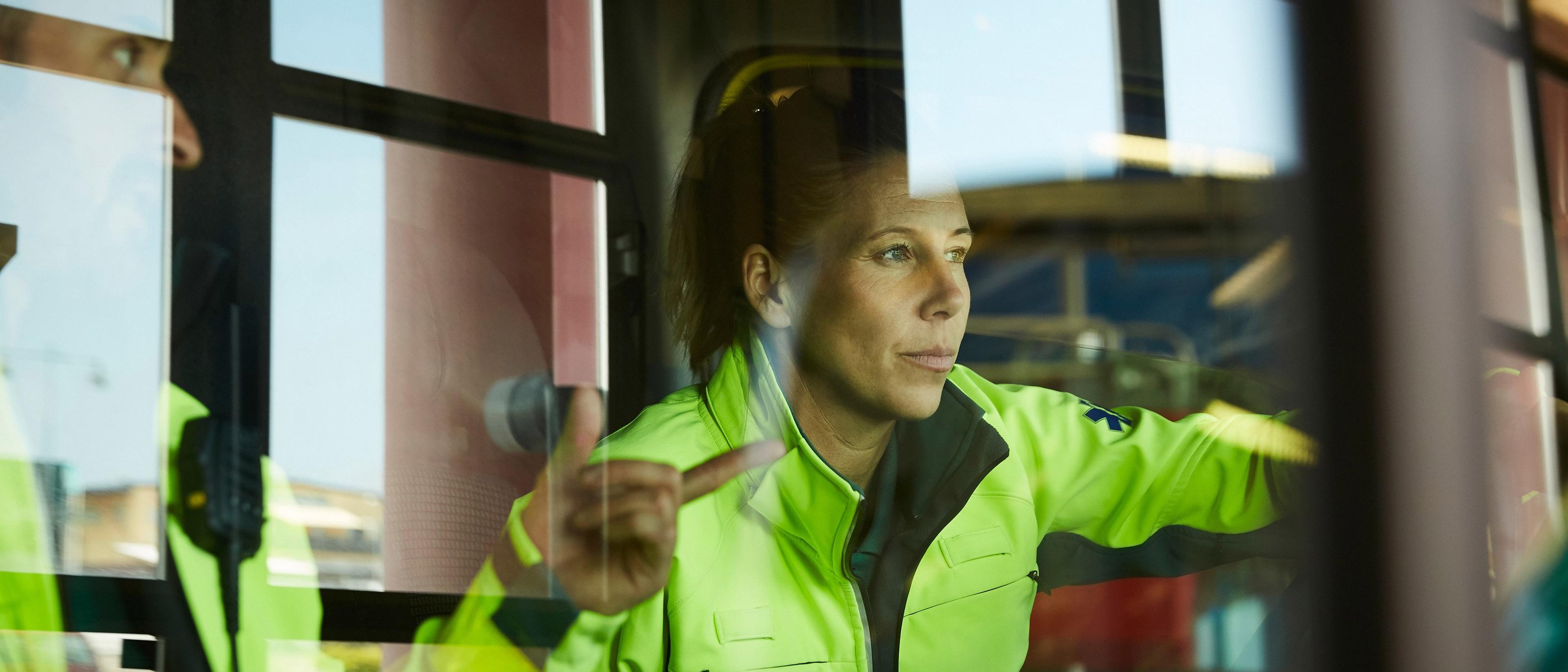 En kvinne i gul refleksjakke sitter inne i et kjøretøy og ser konsentrert ut gjennom frontruten mens hun peker med hånden.
