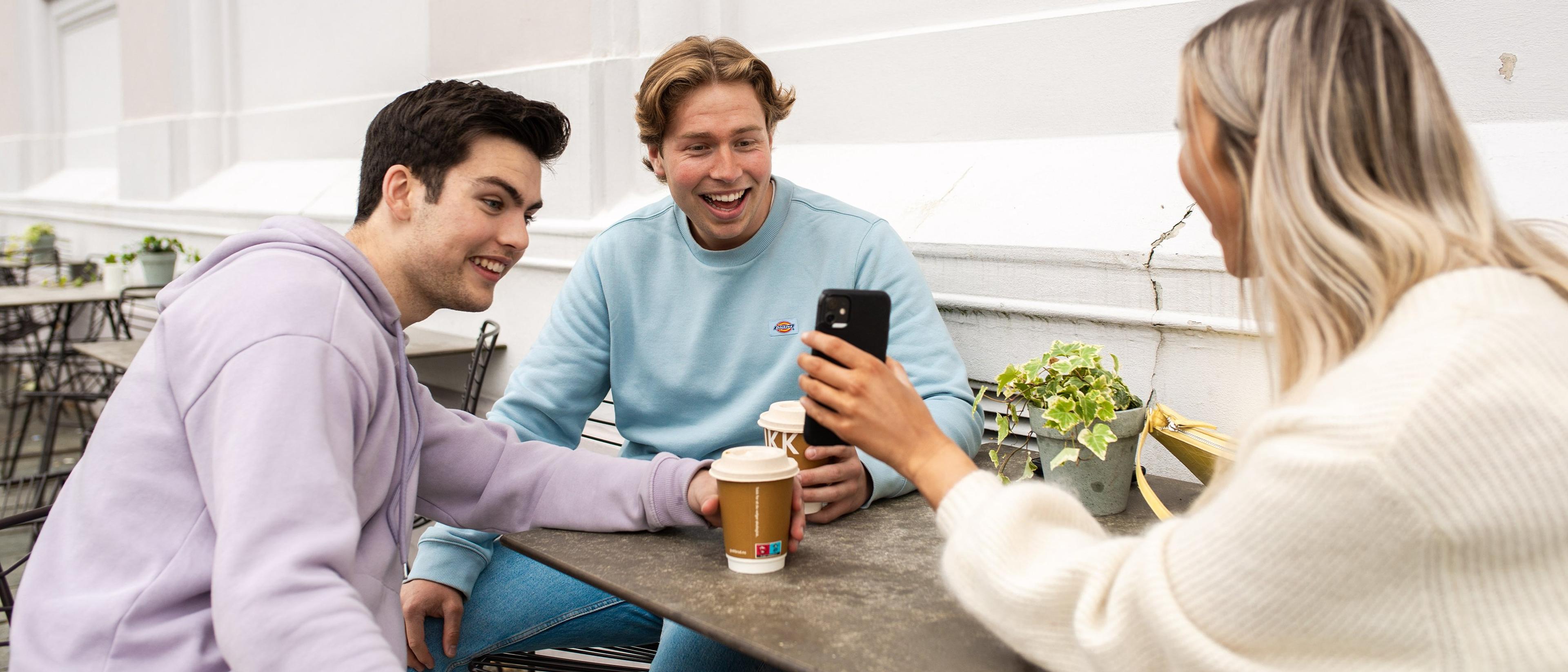 Tre unge voksne sitter rundt et kafébord med kaffekopper og smiler mens kvinnen viser noe på mobiltelefonen sin.