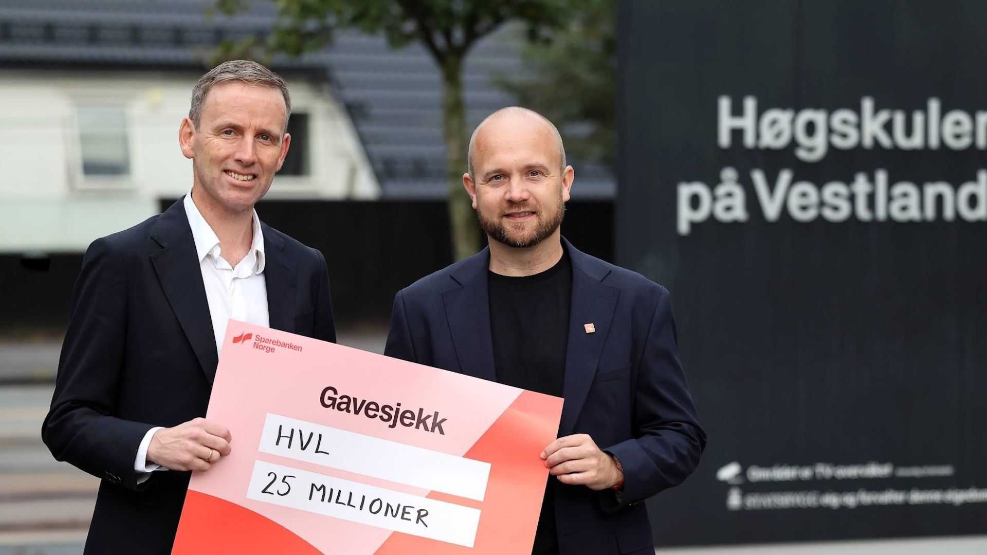 To menn holder en stor gavesjekk på 25 millioner kroner til HVL foran et skilt med teksten «Høgskulen på Vestlandet».