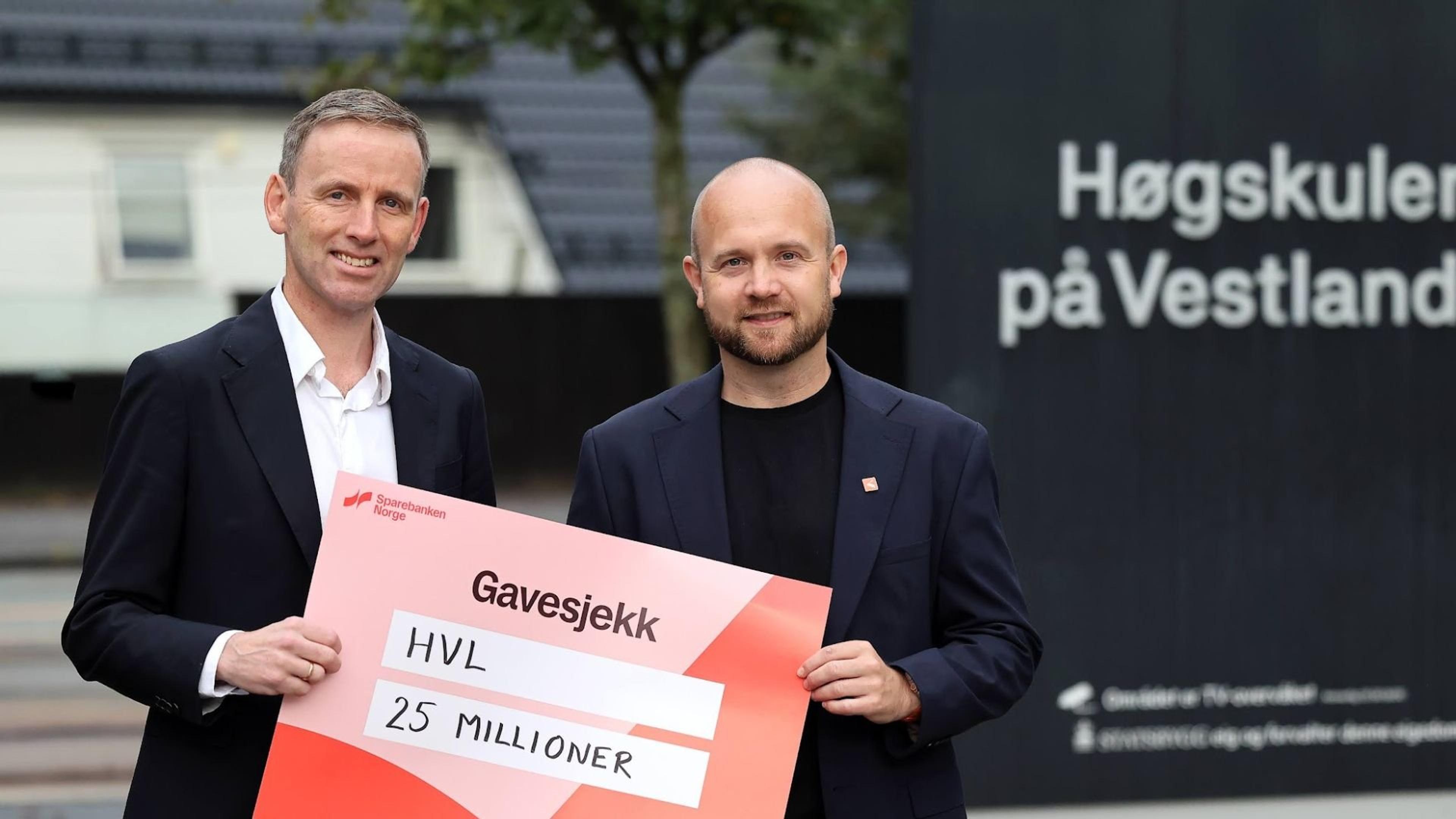 To menn holder en stor gavesjekk på 25 millioner kroner til HVL foran et skilt med teksten «Høgskulen på Vestlandet».